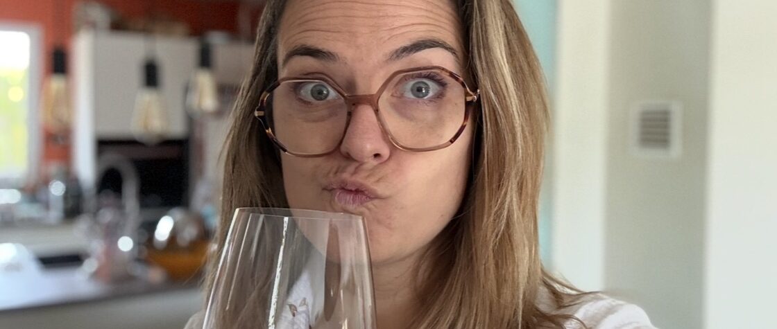Portrait de Charlotte des itinéraires de Charlotte faisant une moue amusée en goûtant un verre de vin rouge dans une cuisine lumineuse, expression qui illustre le fait de ne pas aimer un vin sans se sentir coupable