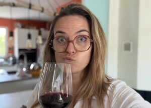 Portrait de Charlotte des itinéraires de Charlotte faisant une moue amusée en goûtant un verre de vin rouge dans une cuisine lumineuse, expression qui illustre le fait de ne pas aimer un vin sans se sentir coupable