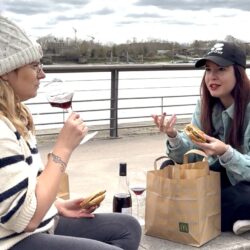 Deux amies assises au bord d’un fleuve, partageant un moment simple avec un verre de vin rouge et un repas improvisé, dans une ambiance d’hiver douce et complice.