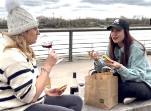 Deux amies assises au bord d’un fleuve, partageant un moment simple avec un verre de vin rouge et un repas improvisé, dans une ambiance d’hiver douce et complice.