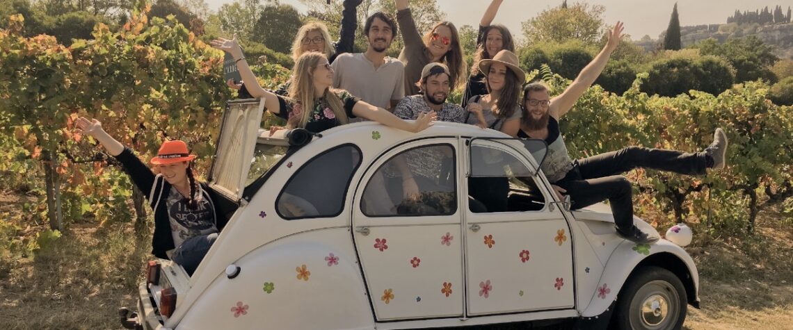Groupe de jeunes voyageurs souriants dans une 2CV fleurie, au milieu des vignes ardéchoises, profitant d’une expérience œnotouristique joyeuse sous la lumière douce de l’automne.