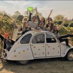 Groupe de jeunes voyageurs souriants dans une 2CV fleurie, au milieu des vignes ardéchoises, profitant d’une expérience œnotouristique joyeuse sous la lumière douce de l’automne.