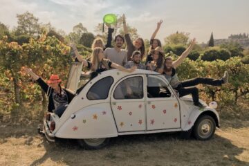 Groupe de jeunes voyageurs souriants dans une 2CV fleurie, au milieu des vignes ardéchoises, profitant d’une expérience œnotouristique joyeuse sous la lumière douce de l’automne.