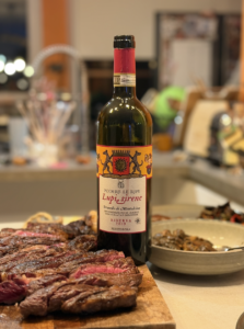 Bouteille de Lupi e Sirene Riserva 2019 de Podere Le Ripi posée sur une table, accompagnée d’entrecôtes grillées et de cèpes