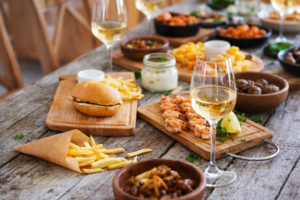 Table conviviale avec plats du quotidien (burger, frites, crevettes) et verres de vin blanc, illustrant des accords mets-vins simples et accessibles.