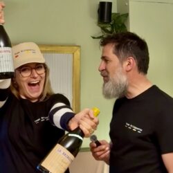 Charlotte souriante levant deux bouteilles de La Bringuette lors du lancement au Georges au Bouscat, aux côtés de Fabien le winemaker, dans une ambiance festive et conviviale.