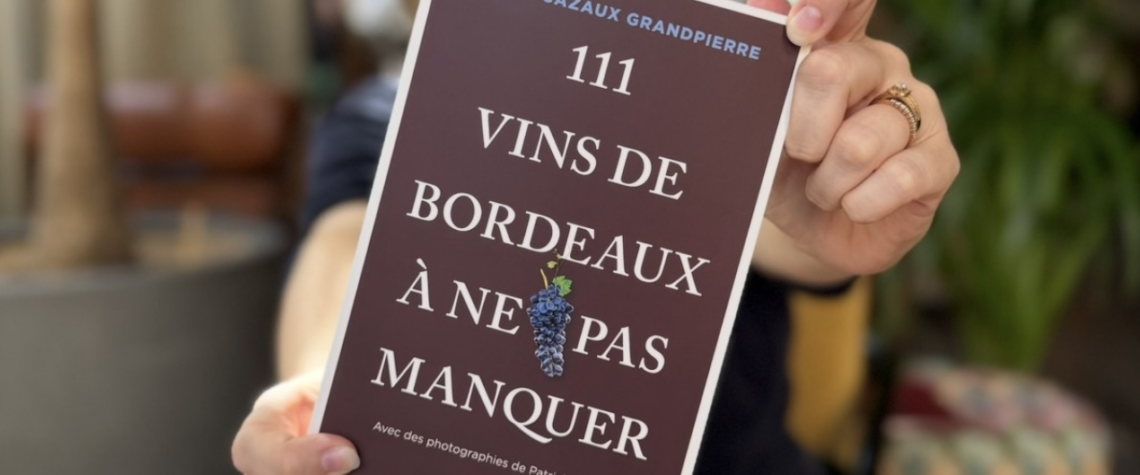 Chloé Cazaux Grandpierre, floue à l’arrière-plan, tend la couverture de son livre 111 vins de Bordeaux à ne pas manquer vers l’objectif.