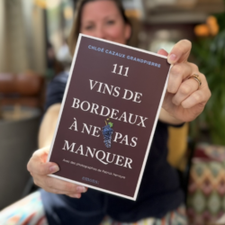 Chloé Cazaux Grandpierre, floue à l’arrière-plan, tend la couverture de son livre 111 vins de Bordeaux à ne pas manquer vers l’objectif.