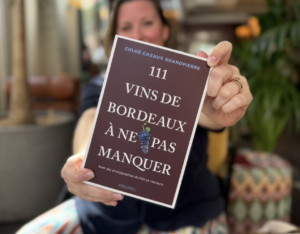 Chloé Cazaux Grandpierre, floue à l’arrière-plan, tend la couverture de son livre 111 vins de Bordeaux à ne pas manquer vers l’objectif.