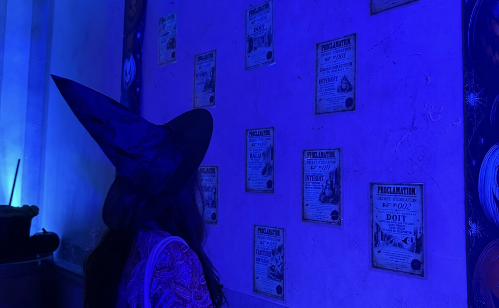 Une apprentie sorcière coiffée d'un grand chapeau pointu découvre les proclamations affichées dans une salle illuminée de bleu et violet au Château Magique de Bordeaux.